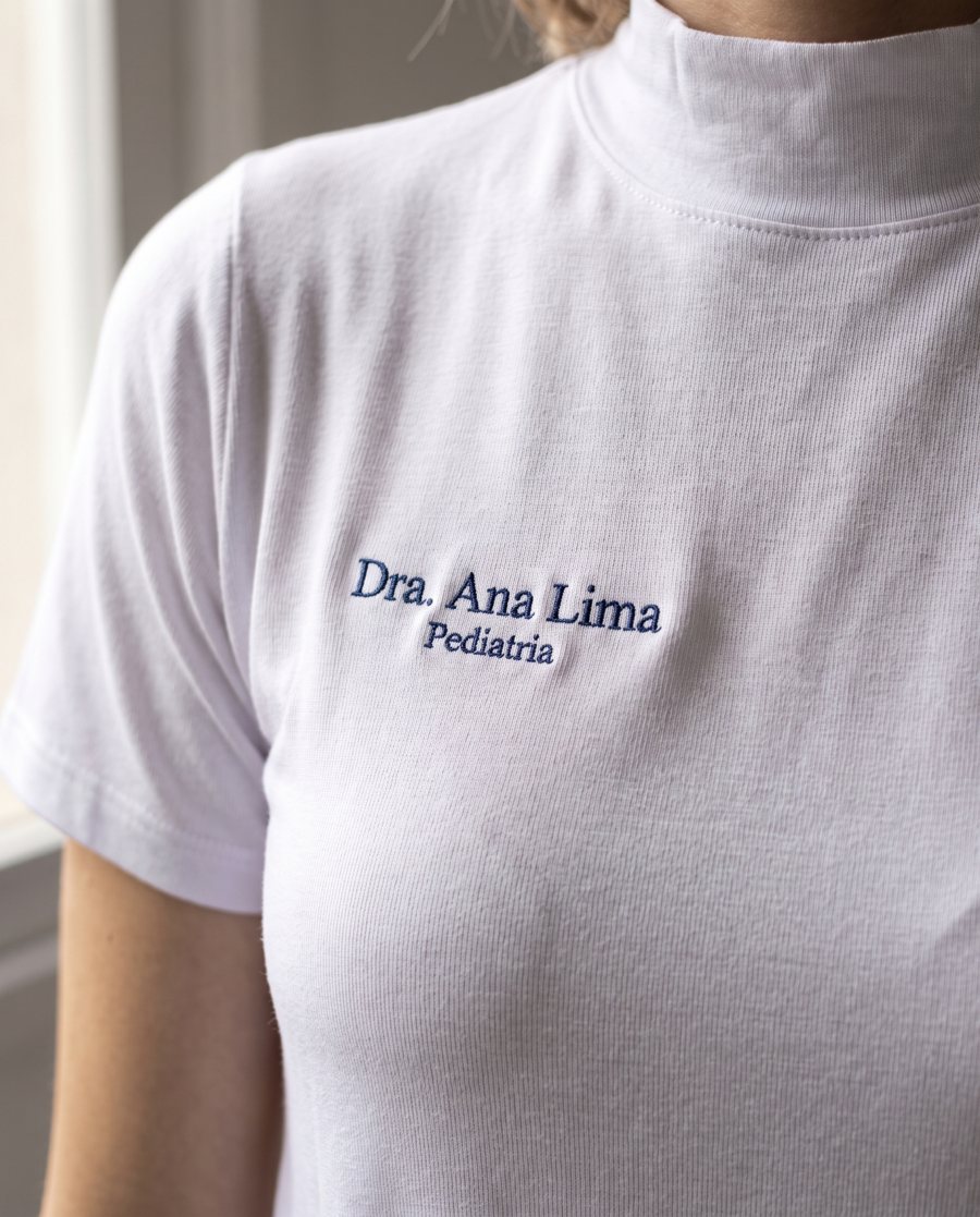 Camiseta branca de gola alta com Dra. Ana Lima e Pediatria bordados em azul-marinho no peito